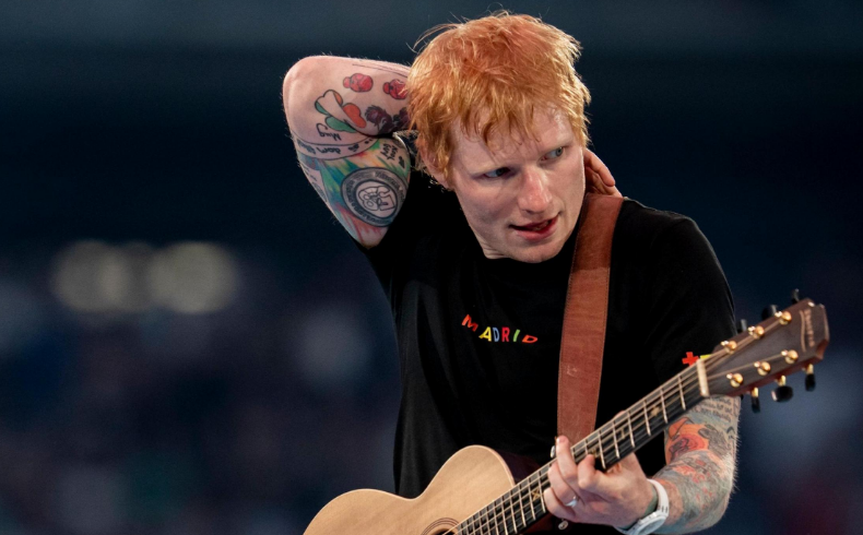 Ed Sheeran smiling and holding his acoustic guitar on a concert stage
