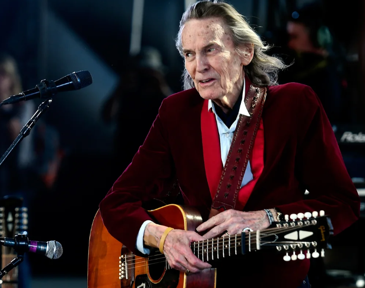 A classic black and white portrait of a young Gordon Lightfoot smiling and playing his acoustic guitar on stage.