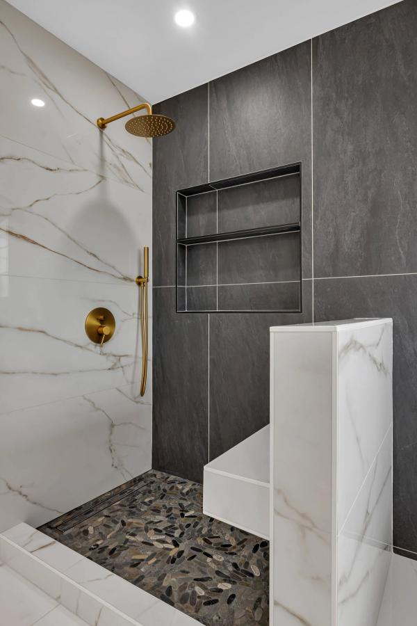 Walk-in shower interior, dark slate tile with dual niches, gold rain head, pebble floor, and marble half-wall