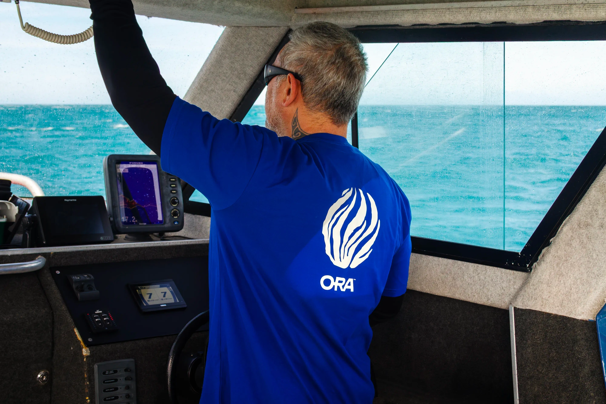 A boat captain pilots his vessel wearing a blue ORA t-shirt with a kelp emblem on the back