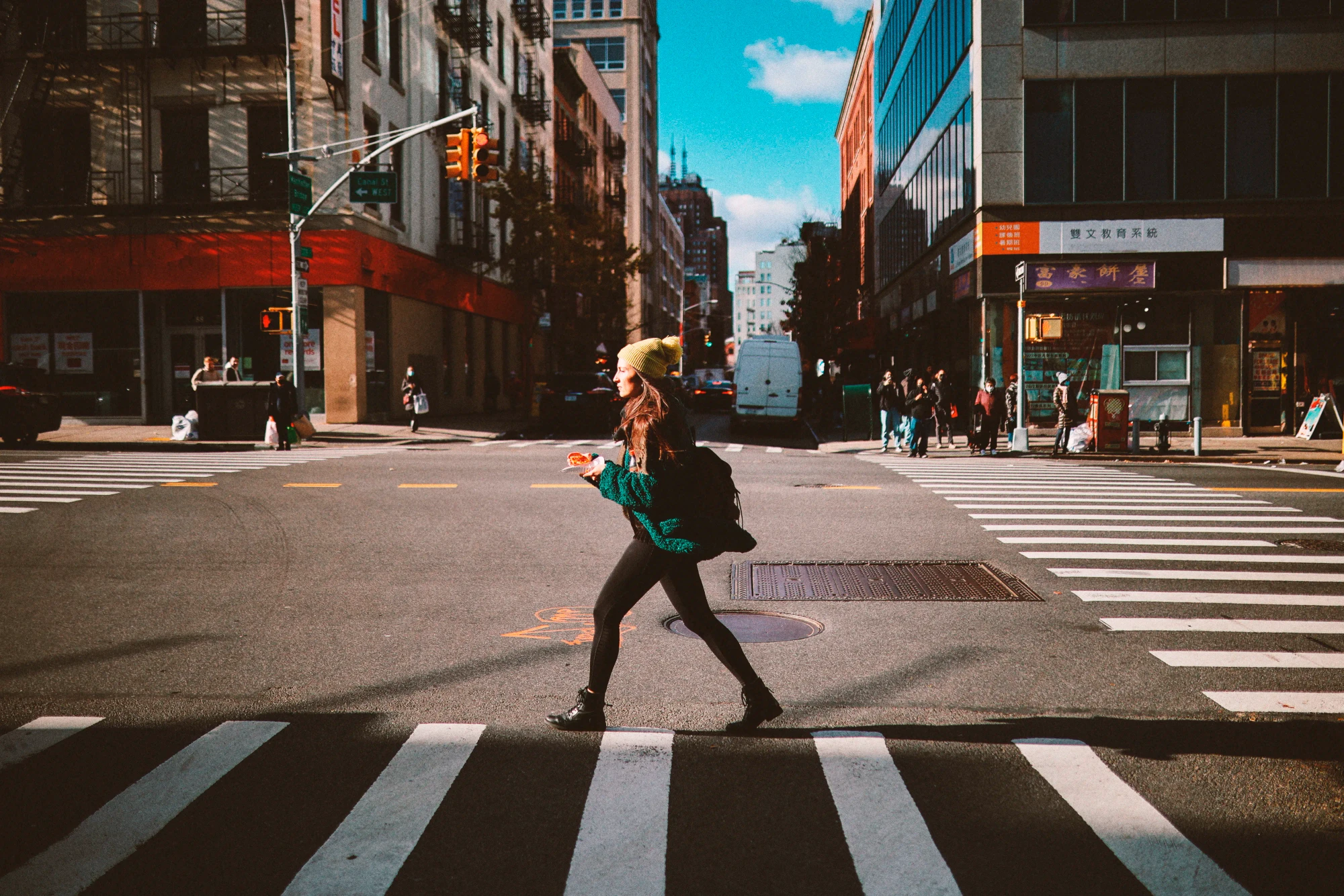 A woman walks across a typical New York intersection, holding a slice of pizza to go.