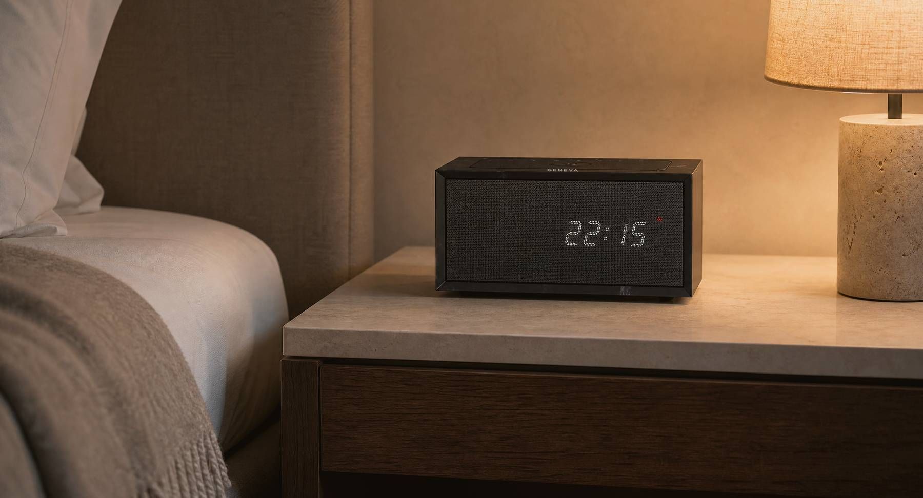 A Geneva Time digital in black on a limestone nightstand beside a bed in the evening