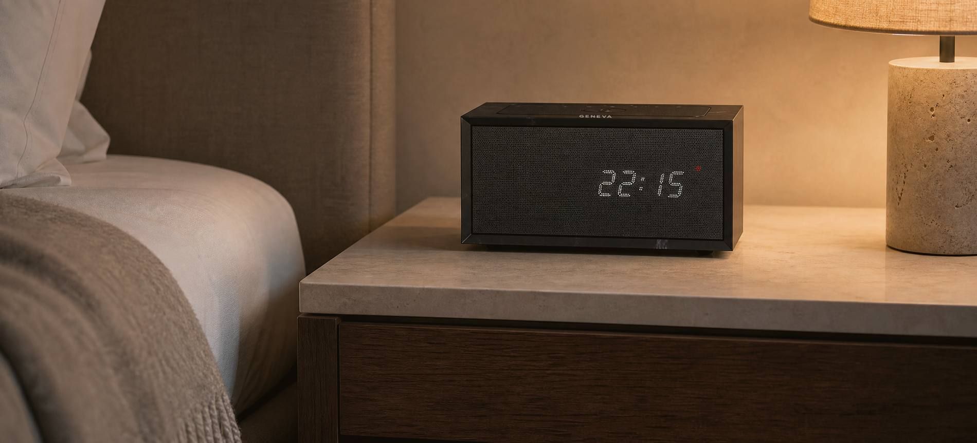 A Geneva Time digital in black on a limestone nightstand beside a bed in a hotel room in the evening