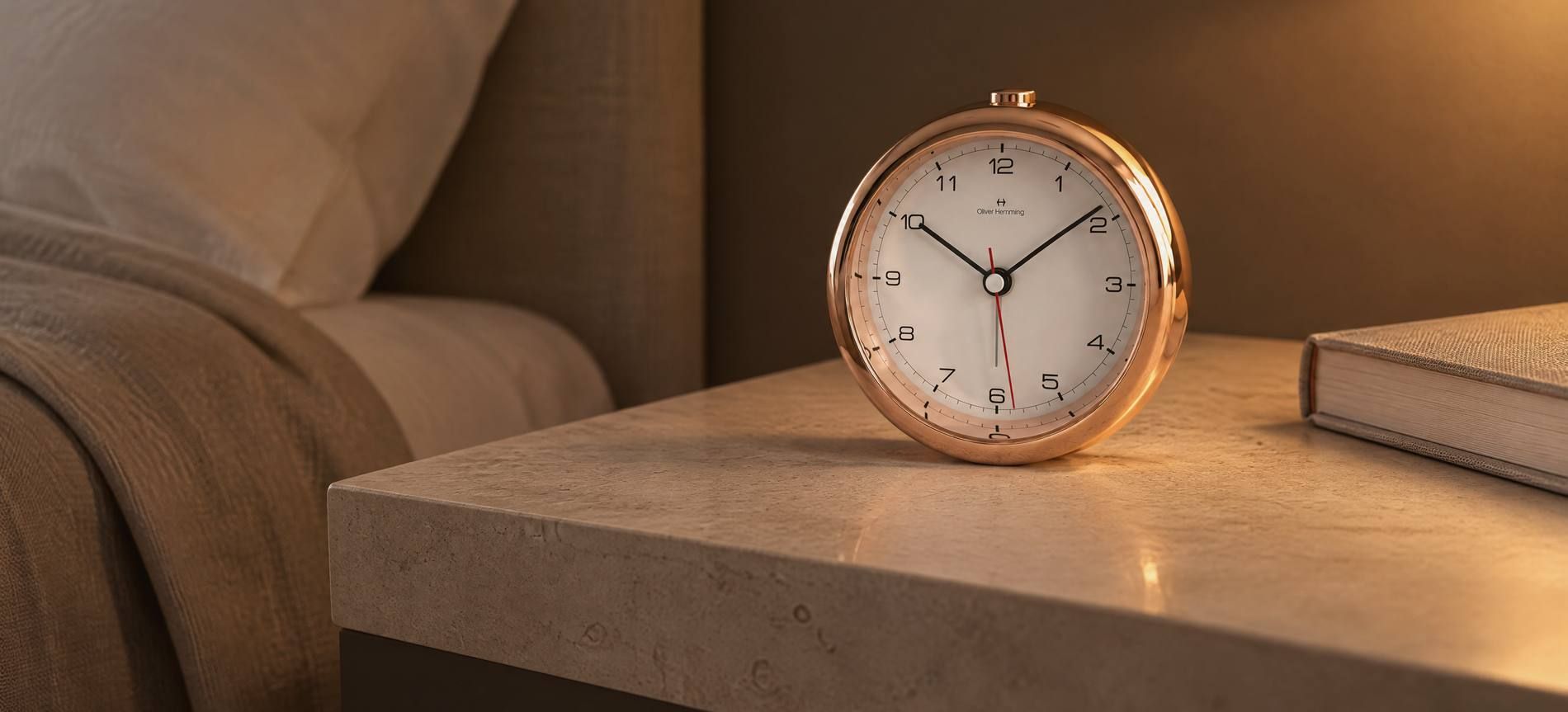 An Oliver Hemming Desire clock in rose gold on a limestone bedside table in a hotel room in the evening