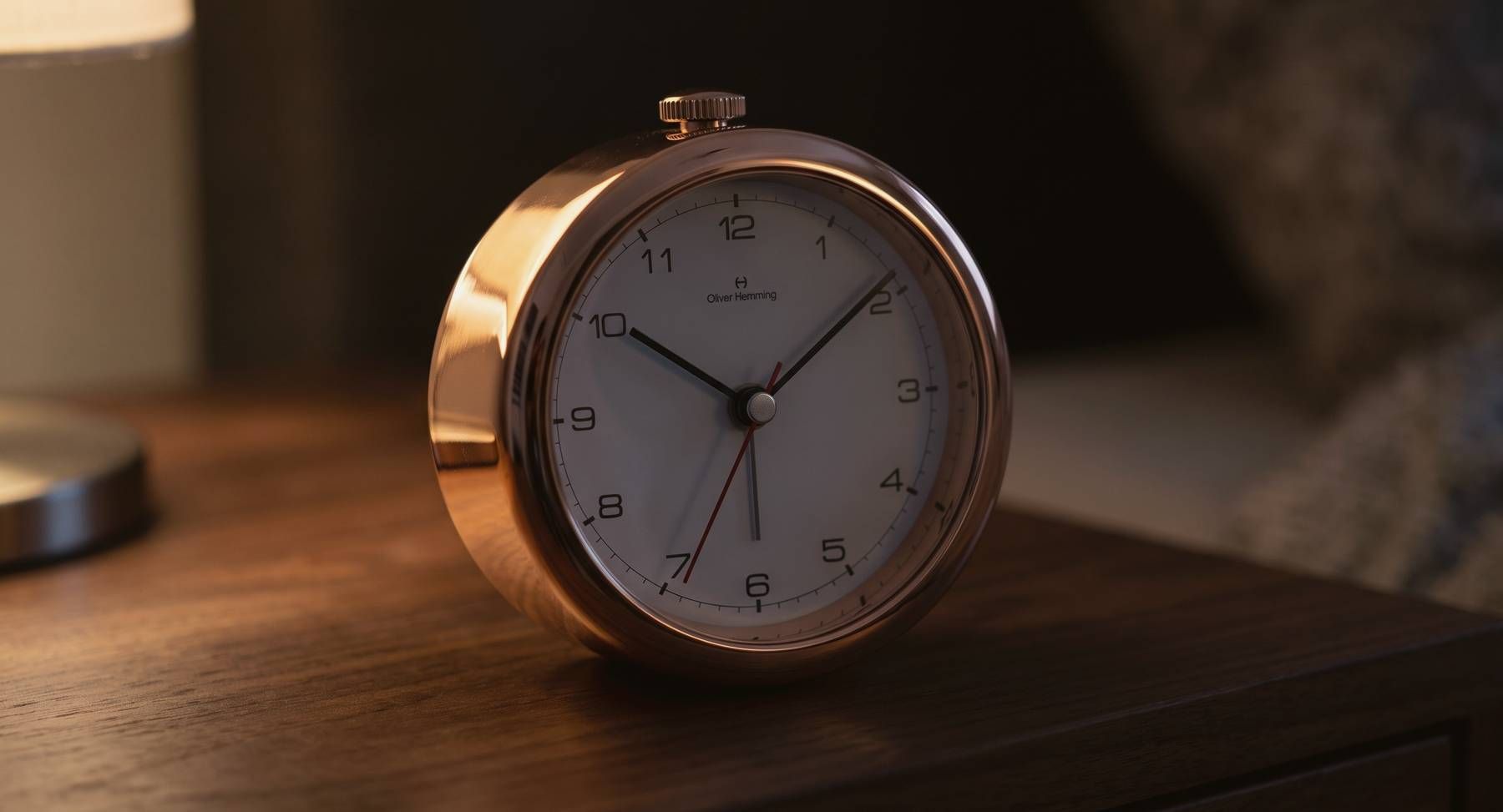 An Oliver Hemming Desire clock in rose gold on a walnut nightstand under lamplight