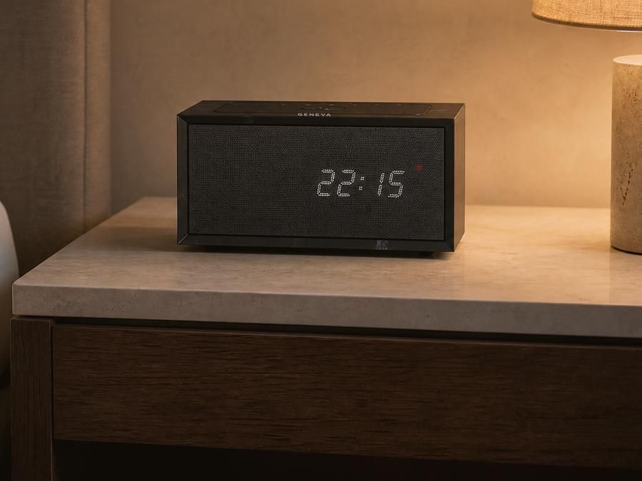 A Geneva Time digital in black on a limestone nightstand beside a bed in a hotel room in the evening