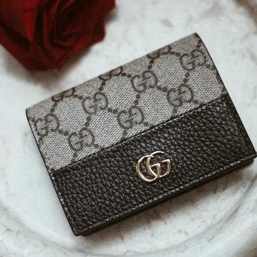 กระเป๋าสตางค์ Gucci ลายโมโนแกรม GG และหนังสีดำเกรน วางบนหินอ่อน มีดอกกุหลาบแดงเบลอๆ ด้านหลัง