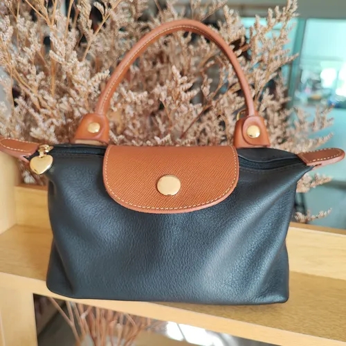 กระเป๋าถือทรงสี่เหลี่ยมคางหมูสีดำ ตัดกับหูหิ้วและฝาปิดหนังสีน้ำตาล วางบนชั้นไม้