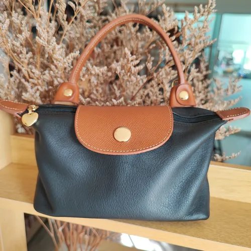 กระเป๋าหนังสีดำขอบน้ำตาล มีหูหิ้วและฝาปิดสีน้ำตาลพร้อมปุ่มทอง วางอยู่หน้าดอกไม้แห้ง