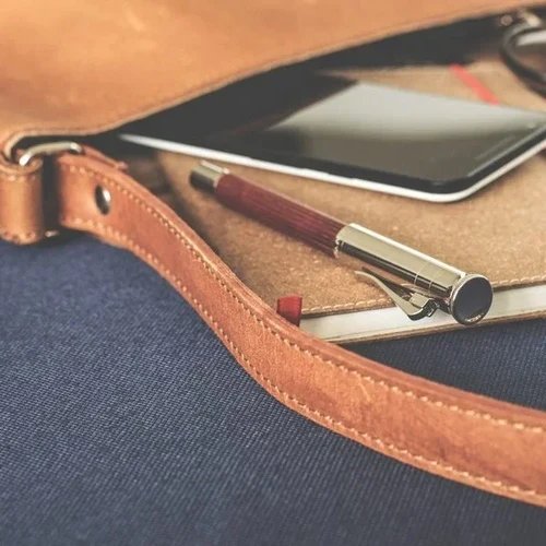 กระเป๋าหนังสีน้ำตาลอ่อนมีสายสะพาย, สมาร์ทโฟน, สมุดโน้ต และปากกา วางบนพื้นผิวสีน้ำเงินเข้ม