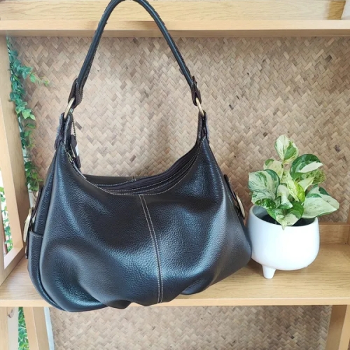 กระเป๋าสะพายหนังแท้สีดำวางอยู่บนชั้นไม้ มีต้นไม้ประดับและฉากหลังสาน