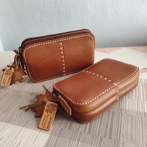 กระเป๋าหนังสีน้ำตาลสองใบวางคู่กัน มีลายเย็บไขว้สีขาวด้านหน้าและพวงกuenุญแจหนังสลักชื่อแบรนด์ 'MARAH'