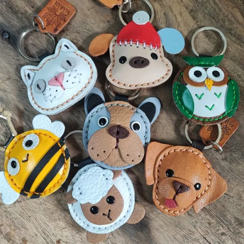 พวงกุญแจหนังแฮนด์เมดรูปหน้าสัตว์ต่างๆ เช่น สุนัข แมว และนกฮูก วางรวมกันบนพื้นผิวไม้