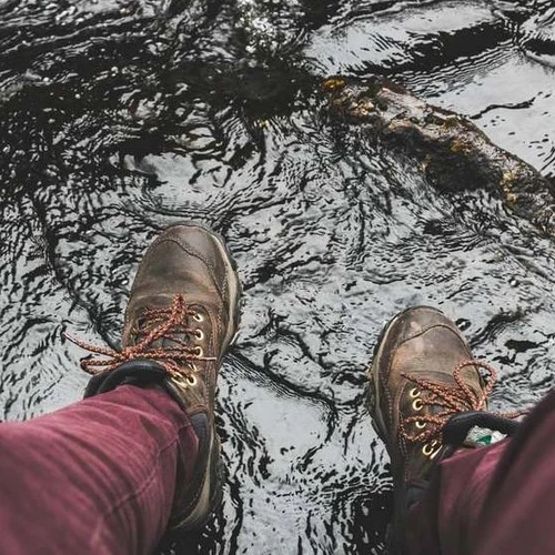 รองเท้าบูทหนังสีน้ำตาลสองข้างกำลังเหยียบอยู่ในน้ำตื้นที่มีคลื่นเล็กน้อย มองเห็นส่วนขากางเกงสีแดงเข้ม