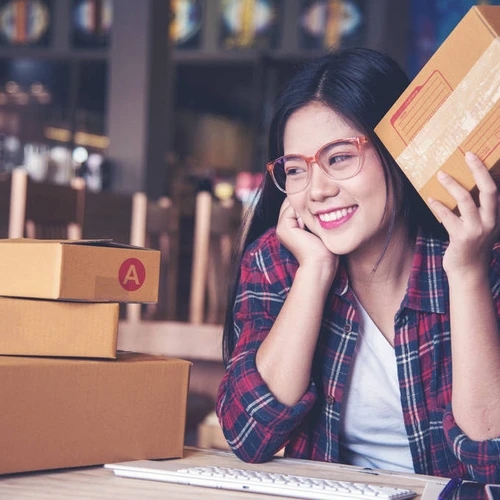 ผู้หญิงเอเชียยิ้มอย่างมีความสุขขณะถือกล่องพัสดุ มีกองกล่องพัสดุอีกหลายใบวางอยู่บนโต๊ะทำงานข้างๆ