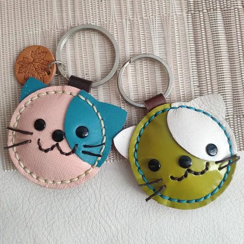 พวงกุญแจหนังรูปแมว 2 ชิ้น สีชมพู-ฟ้า และ สีเขียว-ขาว วางอยู่บนพื้นผิวสีอ่อน แสดงถึงความประณีตและน่ารัก