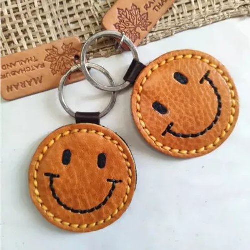 พวงกุญแจหนังรูปหน้ายิ้มสีน้ำตาลสองอันพร้อมห่วงโลหะวางคู่กัน มีป้ายหนังแกะสลักอยู่ด้านหลัง