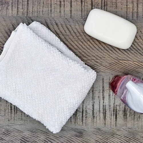 ผ้าขนหนูสีขาว, สบู่ก้อนสีขาว และขวดปั๊มใส่น้ำยาสีแดงวางอยู่บนพื้นไม้