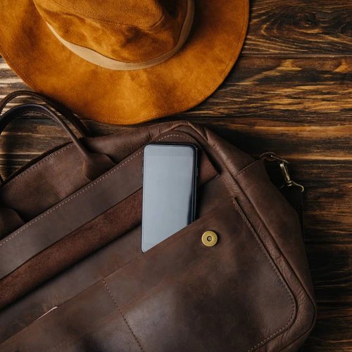 กระเป๋าหนังสีน้ำตาลและหมวกวางบนพื้นไม้ มีโทรศัพท์มือถือเสียบอยู่ในช่องกระเป๋า