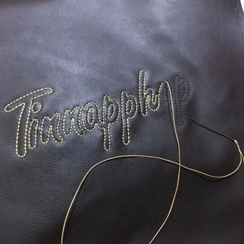 พื้นผิวหนังสีดำที่ปักชื่อ 'TinnaPph' ด้วยด้ายสีเหลืองอ่อน พร้อมเข็มและด้ายที่กำลังปักอยู่