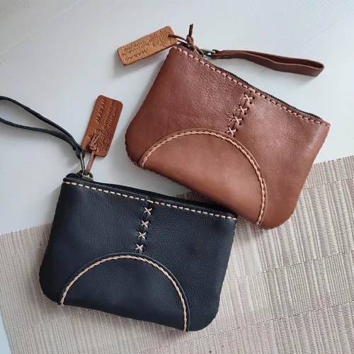 กระเป๋าหนังใบเล็กสองใบสีน้ำตาลและสีดำ วางบนพื้นผิวสีอ่อน พร้อมสายคล้องข้อมือและป้ายหนัง