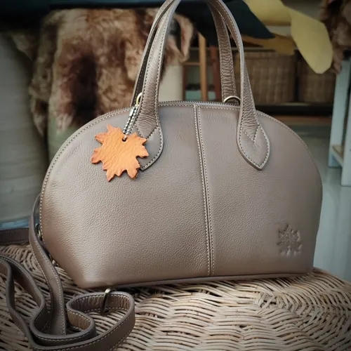 กระเป๋าถือหนังแท้ทรงโดมสีน้ำตาลหม่นพร้อมพวงกุญแจรูปใบไม้ วางอยู่บนตะกร้าหวาย