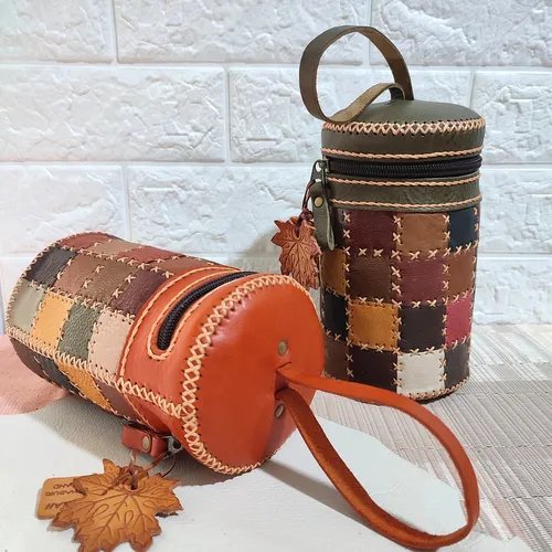 กระเป๋าทรงกระบอกสองใบทำจากหนังวัวเย็บปะติดปะต่อกัน ใบหนึ่งสีส้ม-น้ำตาล อีกใบสีเขียวเข้ม มีงานเย็บมือและจี้รูปใบไม้