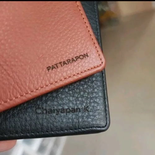 กระเป๋าสตางค์หนังแท้สีน้ำตาลและสีดำสลักชื่อบุคคล เห็นพื้นผิวหนังลายลิ้นจี่ชัดเจน