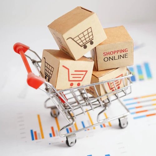 ตะกร้าช้อปปิ้งขนาดเล็กบรรจุกล่องพัสดุ วางอยู่บนกระดาษกราฟแท่ง สื่อถึงการเติบโตของการซื้อขายสินค้าออนไลน์