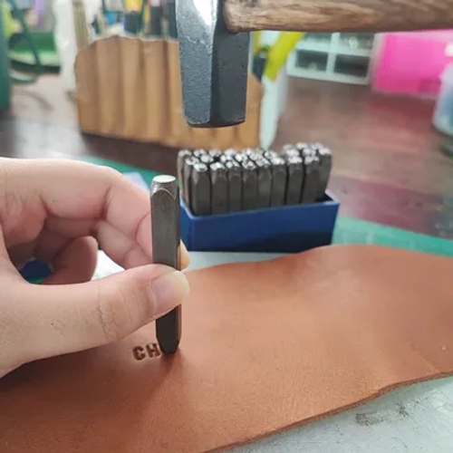 มือตอกตัวอักษร 'H' ลงบนหนังสีน้ำตาล มีค้อนและชุดตัวตอกอยู่ด้านหลัง ตัวอักษร 'CH' ถูกตอกไว้แล้ว