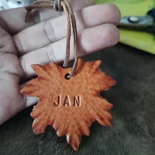 พวงกุญแจหนังสักชื่อรูปใบเมเปิ้ลสีส้มน้ำตาล มีคำว่า 'JAN' สลักอยู่บนผิวหนัง
