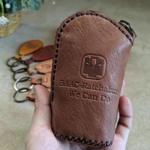กระเป๋าใส่กุญแจหนังสีน้ำตาล สลักโลโก้ BAAC-Ratchaburi และข้อความ 'We Can Do' ถืออยู่ในมือ มีกระเป๋าอื่นอยู่ด้านหลัง