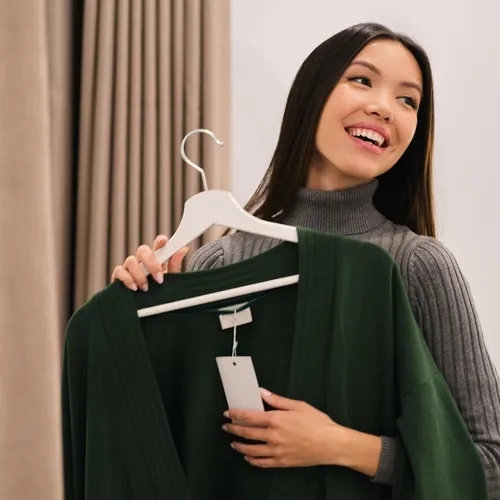 ผู้หญิงยิ้มแย้มถือเสื้อคาร์ดิแกนสีเขียวเข้มพร้อมป้ายราคาบนไม้แขวนเสื้อ