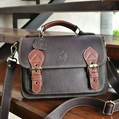 กระเป๋าสะพายหนังสีน้ำตาลเข้ม มีหูหิ้วและสายสะพาย รายละเอียดหัวเข็มขัดสีน้ำตาลอ่อนวางอยู่บนโต๊ะไม้