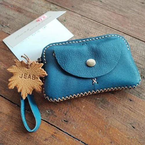 A blue handmade leather coin purse with light stitching, a leaf-shaped 'JEAB' tag, and a greeting card on a wooden surface.