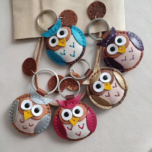 พวงกุญแจหนังสัตว์รูปนกฮูกหลากหลายสี เช่น สีฟ้า, ม่วง, เงิน, แดง และทอง วางเรียงอยู่บนพื้นผิวสีอ่อนพร้อมพวงกุญแจโลหะ