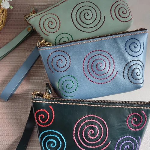 กระเป๋าหนังแฮนด์เมดสามใบสีเขียวมิ้นต์ ฟ้า และเขียวเข้ม/ดำ ประดับด้วยลายปักก้นหอยหลากสีสัน มีสายคล้องข้อมือและซิป