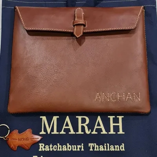 กระเป๋าหนังสีน้ำตาลเข้มพร้อมชื่อ 'ANCHAN' สลักอยู่ด้านหน้า และพวงกุญแจหนังรูปใบไม้ที่มีคำว่า 'NONG' วางอยู่บนถุงสีน้ำเงิน