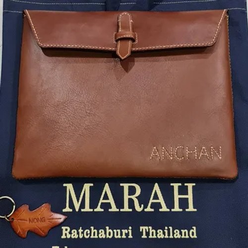 กระเป๋าหนังสีน้ำตาลเข้มพร้อมชื่อ 'ANCHAN' สลักอยู่ด้านหน้า และพวงกุญแจหนังรูปใบไม้ที่มีคำว่า 'NONG' วางอยู่บนถุงสีน้ำเงิน