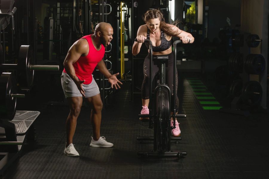 Personal trainer shouting encouragement as a woman pushes hard on an air bike in a dark, moody gym, Shelly Beach.