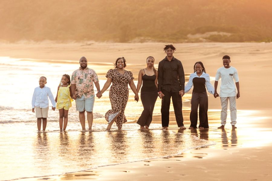 Large family group holding hands and walking together through shallow waves on the beach at golden hour.