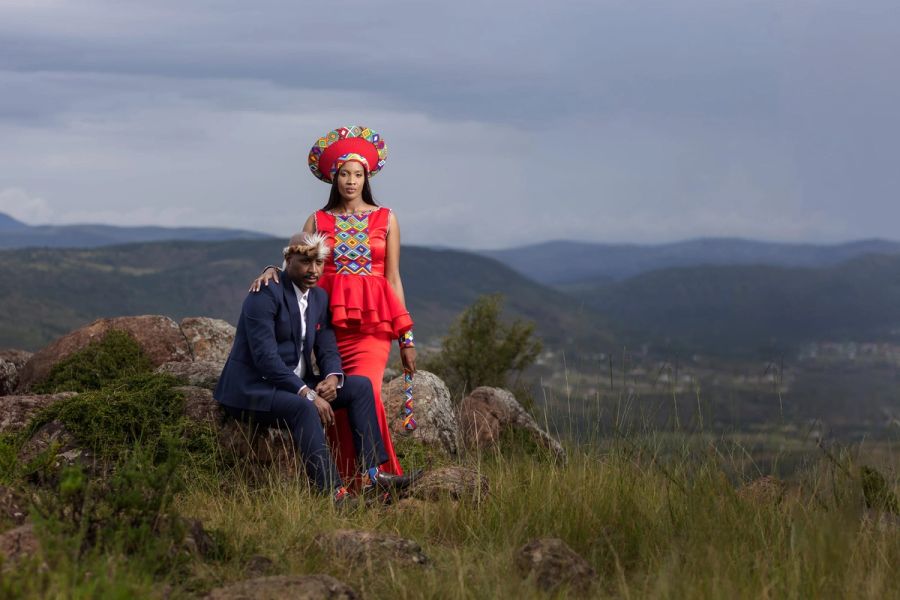 Couple in traditional attire, the woman in a red isicholo headdress and red dress, posing on rocks above a green valley, Ntabankulu.