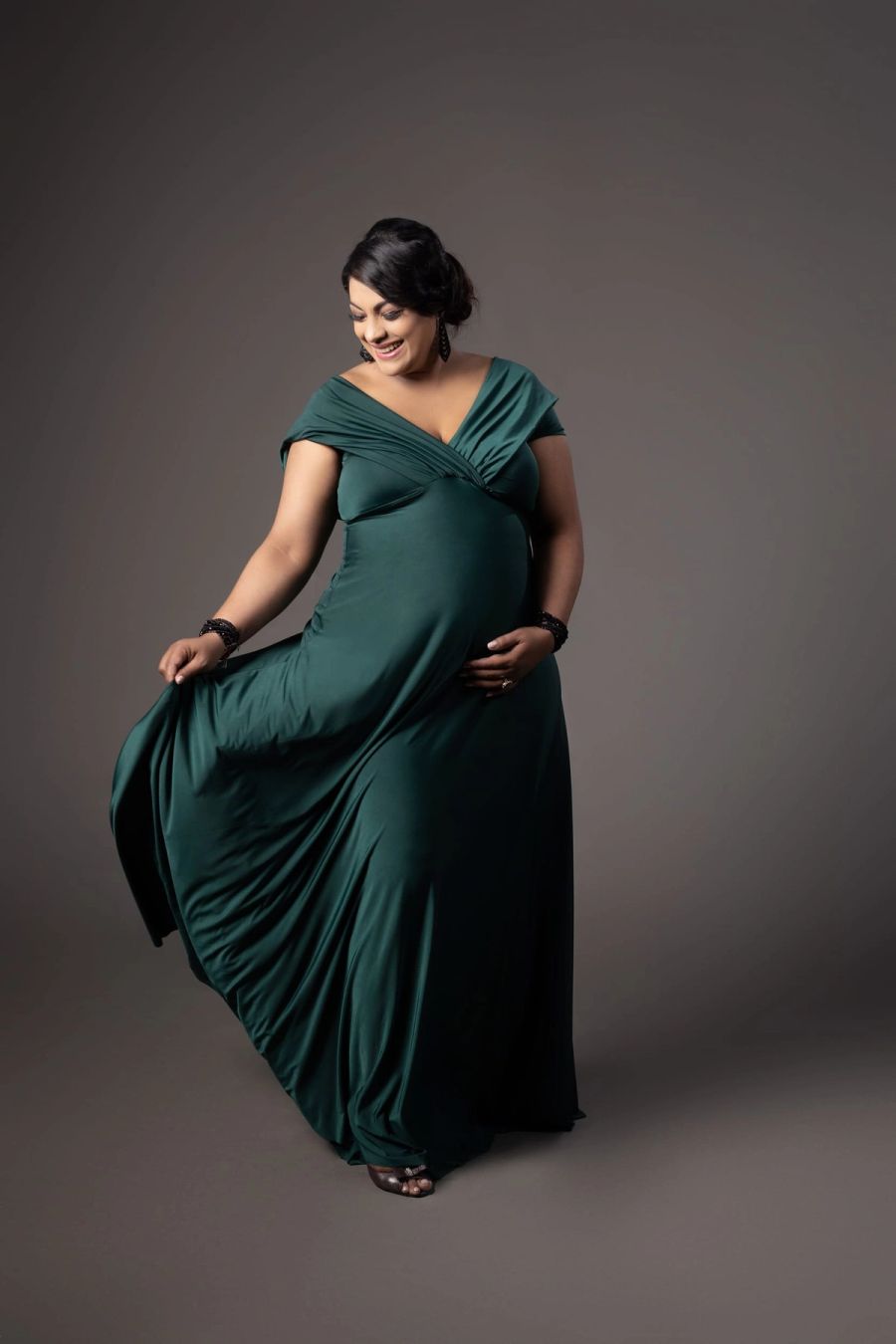 Expectant mother in a flowing emerald green gown, cradling her bump, against a grey studio backdrop, Studio 039, Sea Park.