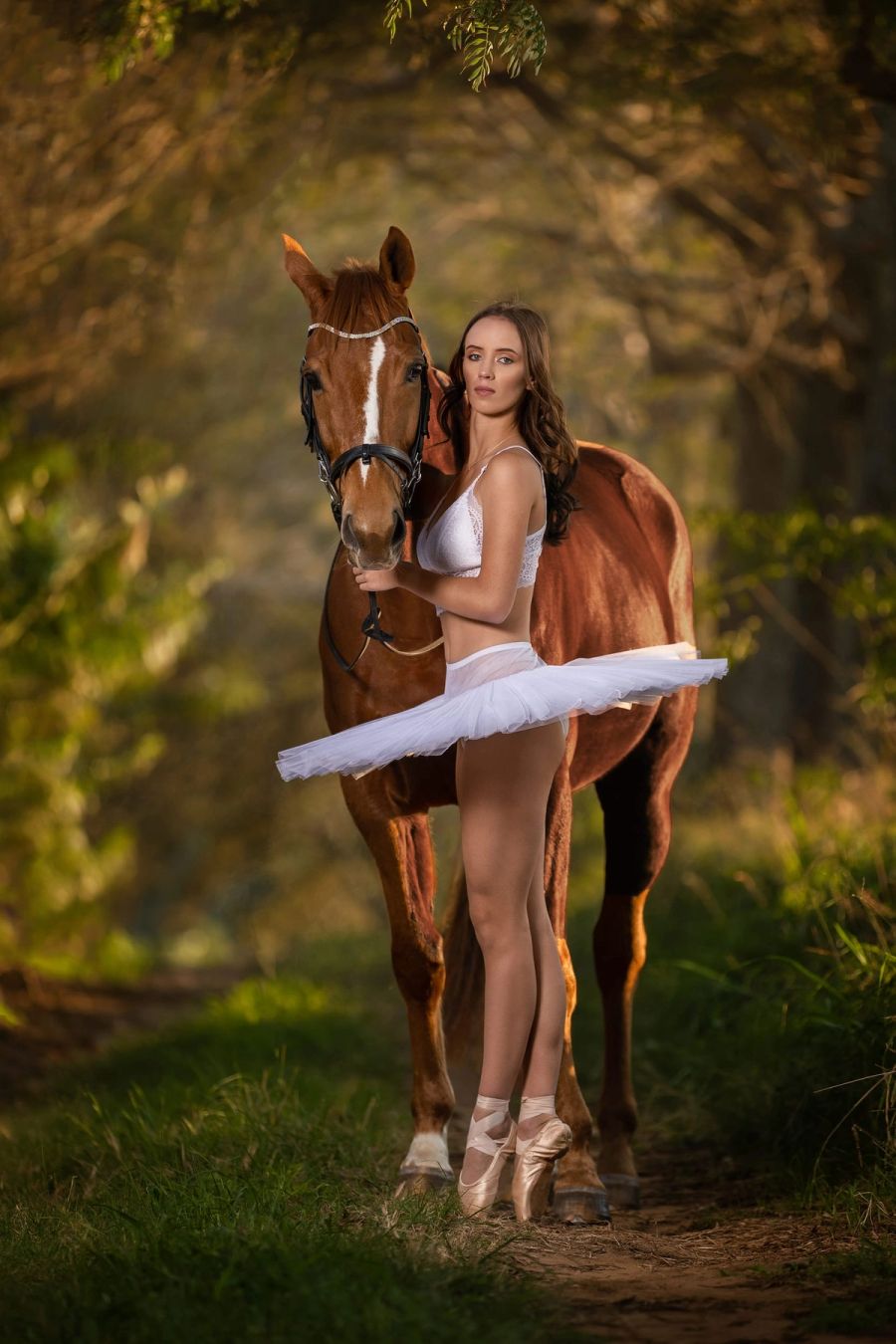 Ballerina in a white tutu and pointe shoes standing beside a chestnut horse on a forest path in warm evening light, Southbroom.