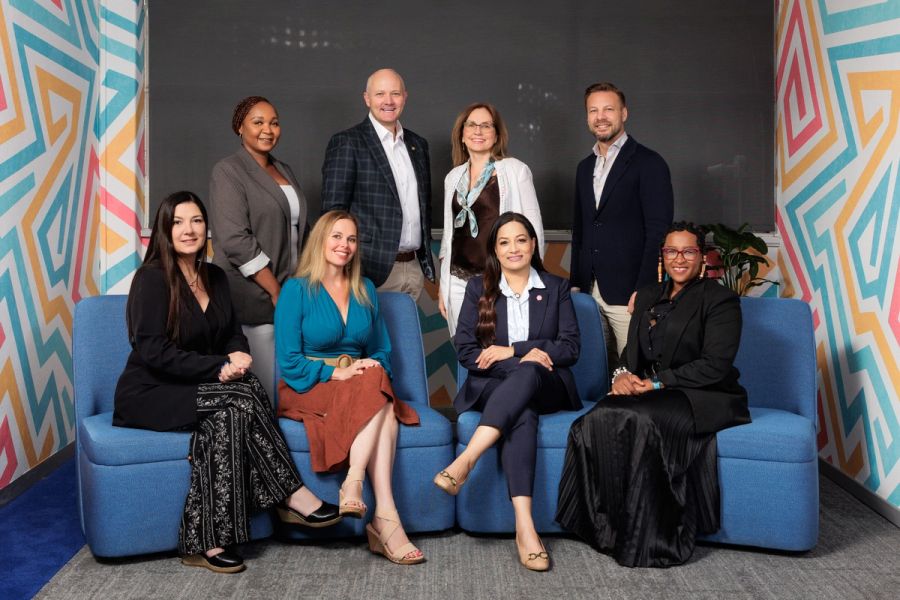 Corporate team of eight colleagues posing together on blue chairs in a modern meeting room with colourful geometric wall art.