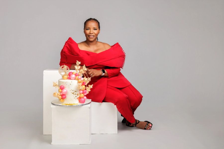 Woman in a red outfit seated beside an elegant decorated birthday cake, studio backdrop.