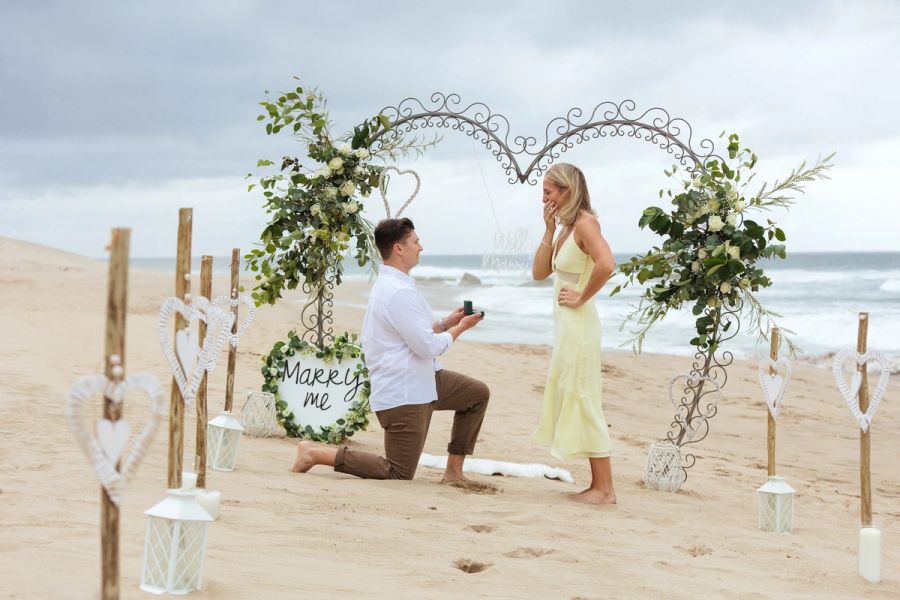 Man kneeling on the beach proposing with a ring to a woman under a heart-shaped floral arch reading 'Marry Me'.