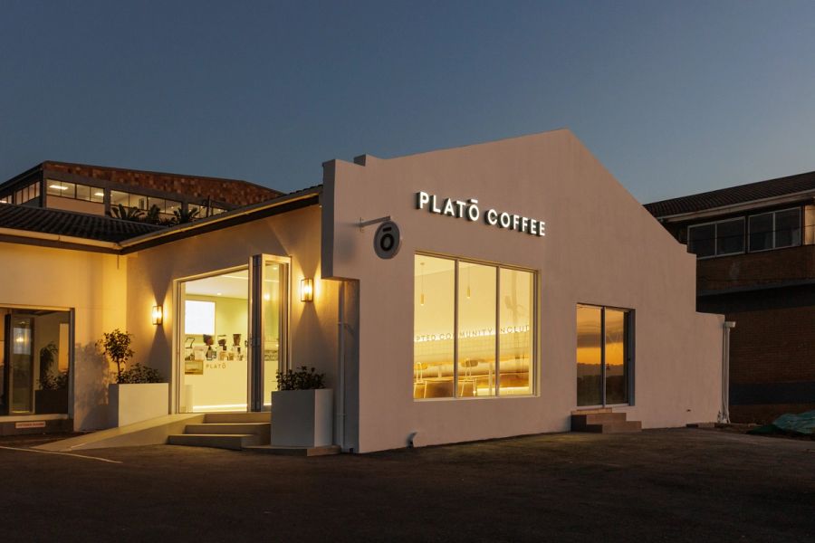 Exterior of a Plato Coffee store at dusk, with warm light glowing through the large front windows.