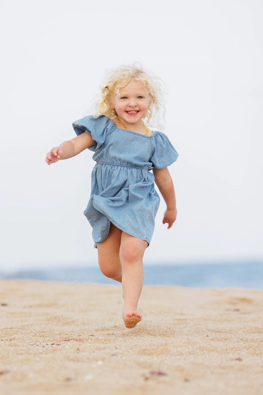 Little girl with blonde curls running barefoot across the sand in a denim dress, St Michael’s Beach.