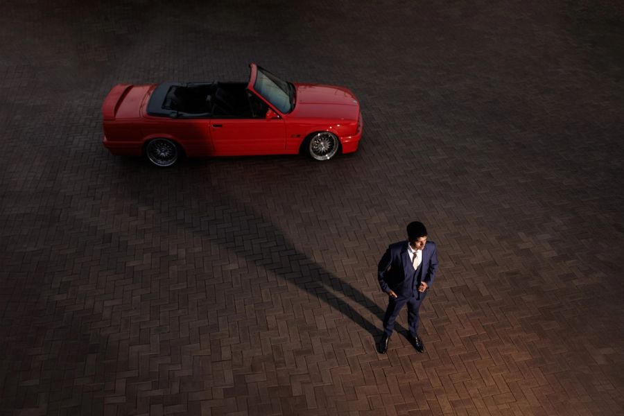Aerial view of a teenager in a formal suit standing beside a red convertible car on a paved driveway.